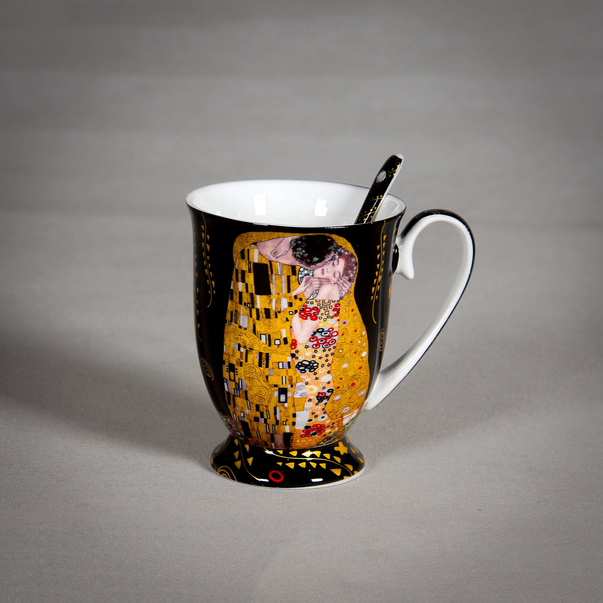 A porcelain mug with a handle and matching spoon, featuring Gustav Klimt's famous painting "The Kiss" in gold, black, and colorful accents on a black background. The mug is set against a plain gray surface.